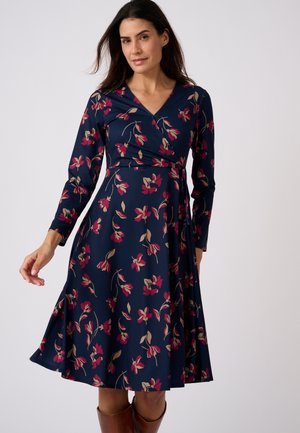 Vrouw draagt een marineblauwe bloemenprint lange mouwen wikkeljurk en staat tegen een effen lichte achtergrond.
