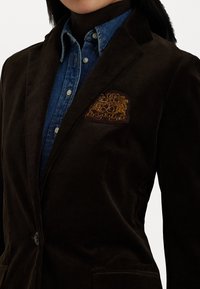 Καφέ καμουφλάζ σακάκι με αυλακωτό γιακά, με διακριτική κεντημένη ταυτότητα σε μπεζ και φορεμένο πάνω από ένα μπλε τζην πουκάμισο.