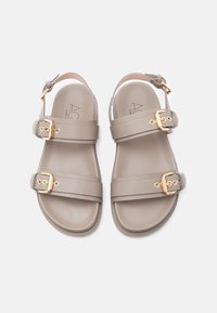 Ζευγάρι σανδάλια σε απόχρωση ταούπε με δύο χρυσά λουράκια με αγκράφες και λουράκι αστραγάλου σε λευκό φόντο.