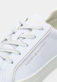 Balti odiniai sportbačiai su plokščiomis raišteliais, apvaliu nosiu ir auksiniu "TOMMY HILFIGER" prekės ženklu šone. Matomas tekstūruotas pado detalės dizainas.