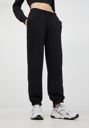 Pantaloni sportivi neri con cintura elastica e polsini, dotati di tasche laterali. Abbinati a scarpe da ginnastica bianche. Texture liscia, simile al cotone.