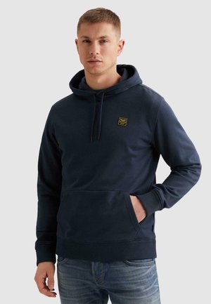 Mann trägt einen marineblauen Kapuzenpullover mit Fronttasche und kleinem gelbem Logo, steht mit einer Hand in der Tasche vor einem einfarbigen Hintergrund.