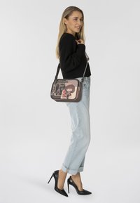 Borsa a spalla con un design illustrato in rosa e marrone, caratterizzato da personaggi da cartone animato e testo. Il materiale è liscio; ha una lunga tracolla regolabile.