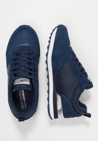 Ett par marinblå Skechers sneakers med mesh-tyg, mockadetaljer, vita sulor och silveraccenter mot en vit bakgrund.
