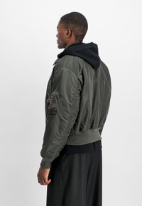 Green bomber jacket made of shiny nylon with ribbed cuffs and hem, featuring side pockets and a hooded black sweatshirt underneath.