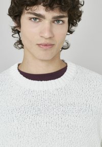 Pull en maille blanche avec un motif texturé, doté d'un col rond côtelé et d'un col intérieur violet. Porté par une personne aux cheveux ondulés.