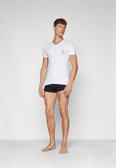 Bela kratka majica s kratkimi rokavi in okroglo ovratnikom ter majhnim logotipom, kombinirana s črnimi kratkimi hlačami; model stoji bosa na nevtralnem ozadju.