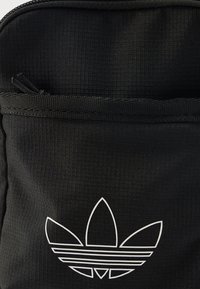 Črn nahrbtnik iz tkanine z teksturirano površino in belim logotipom Adidas z tremi listnim podobnimi podaljški in vzporednimi črtami.