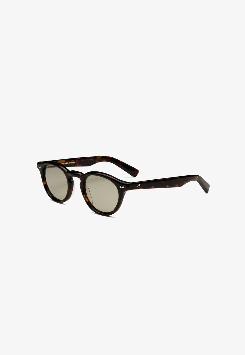 Lunettes de soleil à monture ronde en écaille avec verres teintés foncés et pont en forme de trou de serrure, inclinées sur fond blanc.