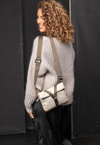 Lichtgrijze messenger tas met een overslagklep, donkergroene bandjes en beige accenten. Gebreide trui en zwarte rok gedragen door een model.