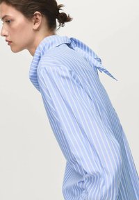 Young woman with brown hair in a bun wearing a blue and white striped shirt with a neck tie detail, facing left on a plain background.