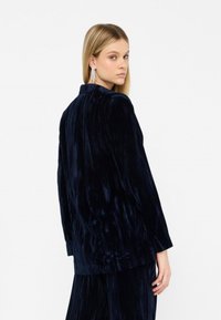 Blazer de terciopelo azul marino con un cuello sencillo, mangas largas y textura vertical; modelo de pie ligeramente de espaldas, luciendo pendientes colgantes.