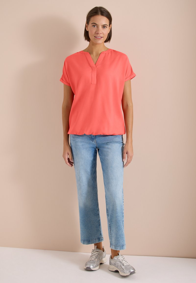 Femme debout de face, portant un haut corail à manches courtes, un jean bleu clair et des baskets argentées, sur un fond beige uni.