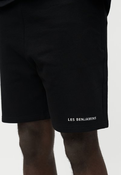 Μαύρο σορτς που φοριέται από άτομο με σκουρόχρωμο δέρμα, με λευκό λογότυπο κειμένου "LES BENJAMINS" κοντά στο τελείωμα στο αριστερό πόδι.