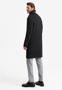 Abrigo de lana negro con un cuello alto y un corte entallado, combinado con pantalones grises con estampado y botas de cuero negras.