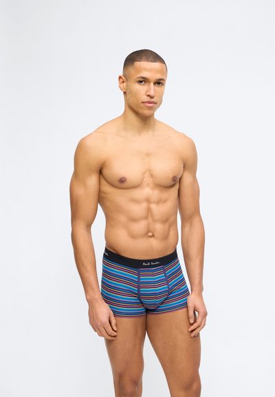 Μυώδης άνδρας στέκεται φορώντας μπλε μποξεράκια με οριζόντιες πολύχρωμες ρίγες και μαύρη ζώνη μέσης, μπροστά σε απλό φωτεινό φόντο.