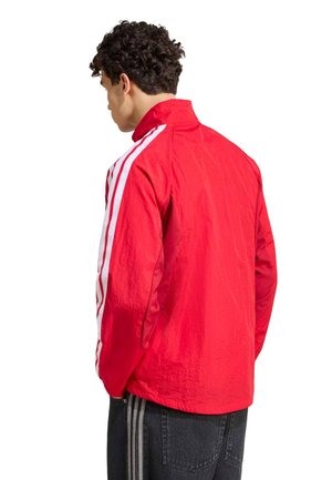 Hombre con cabello rizado que lleva una chaqueta roja con rayas blancas en las mangas, de espaldas sobre un fondo blanco.