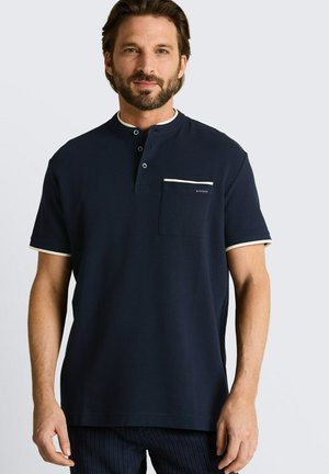 Man met kort bruin haar en baard die een marineblauw kortemouwenoverhemd draagt met een zak en witte bies aan kraag en mouwen.