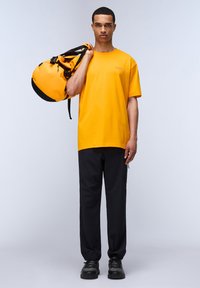 Žluté bavlněné tričko, černé kalhoty a černé tenisky. Model drží oranžovou sportovní tašku s černými popruhy, která má kulatý tvar a zip.