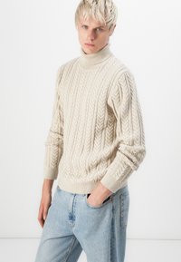 Il pullover beige a collo alto con lavorazione a trecce presenta polsini e orlo a coste, un motivo testurizzato e una vestibilità morbida. Abbinato a jeans azzurri chiari.