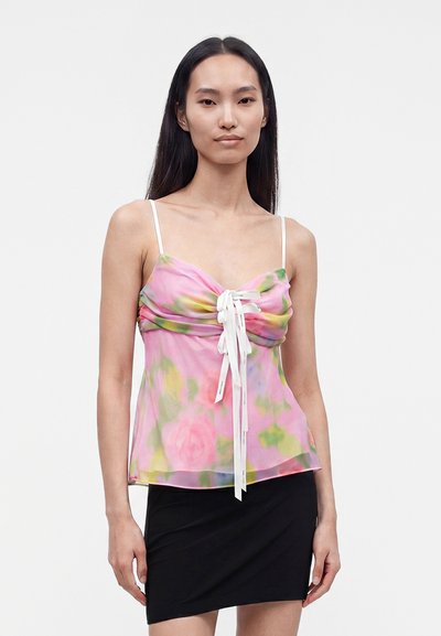 Femme aux longs cheveux lisses portant un haut sans manches fleuri rose et vert avec des liens blancs et une jupe noire courte, face à l'avant, expression neutre.