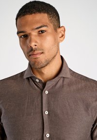 Bruine overhem shirt met een ontspannen kraag, voorzien van witte knopen en een gestructureerde stof, gedragen door een man met kort haar.