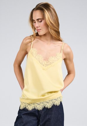 Kvinde iført en lysegul camisole med blonder og mørke bukser, kigger ned med hænderne i lommerne mod en ensfarvet baggrund.