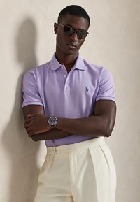 Homme portant un polo violet, un pantalon plissé crème, des lunettes de soleil et une montre-bracelet, debout, les bras croisés contre un mur beige.