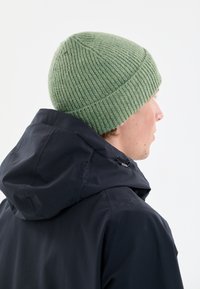 Gröna ribbade stickade mössan med rundad krön och avsmalnande passform, bärs över en mörk huva jacka som visar upp den strukturerade tyget.