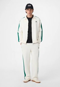 White tracksuit with green side stripes, zip-up jacket, and matching pants, paired with a black cap and white shoes.