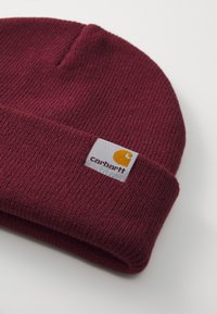 Burgundy knitted beanie with a folded cuff, featuring a square white label with a yellow logo and text. Soft texture with ribbed design.