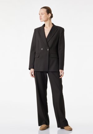 Kobieta w czarnej, dwurzędowej marynarce i dopasowanych szerokich spodniach, stojąca z rozluźnionymi rękami, patrząca w lewo na białym tle.