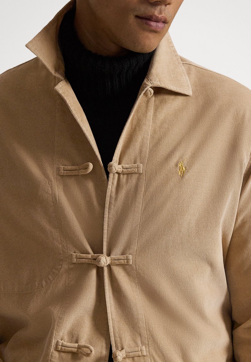 Mann trägt eine beige Cordjacke mit Knebelknöpfen und einen schwarzen Rollkragenpullover darunter, zeigt Oberkörper und Hals.