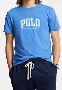 Blå bomullströja med "POLO RALPH LAUREN" i vit text. Kort ärm, rund halsringning. Bärs med mörka träningsoverallsbyxor och vit snörning.