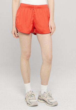 Persona che indossa pantaloncini sportivi arancione brillante, calzini bianchi a metà polpaccio, scarpe da ginnastica beige e grigie, in piedi contro uno sfondo chiaro e semplice.