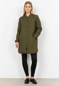 Olijfgroene gewatteerde jas met een rits, geribbelde kraag en zijzakken, gecombineerd met zwarte slim-fit broek en loafers.