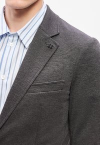 Γκρι σακάκι με υφή υφάσματος, πέτο με σχισμή και μία τσέπη στο στήθος. Κάτω από αυτό, ένα ανοιχτό μπλε ριγωτό πουκάμισο με κουμπιά.