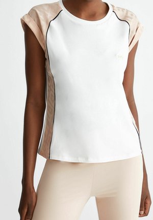 Kobieta ubrana w biały top bez rękawów z beżowymi wzorzystymi panelami po bokach i beżowe dopasowane spodnie, stojąca na jednolitym tle.