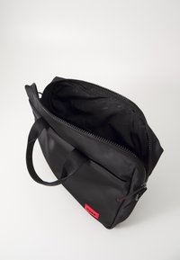 Sort nylon taske med to håndtag, lynlås lukning og rød logo tag. Indersiden er foret med sort stof og har et mønster af logoer.