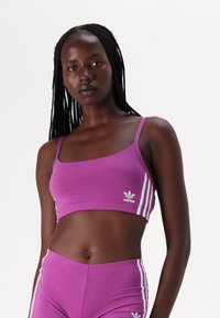 Violet sportinis topas su plonomis petnešomis. Jame yra baltas Adidas logotipas ir trys baltos juostelės šone. Sklandus audinys, prigludęs dizainas.