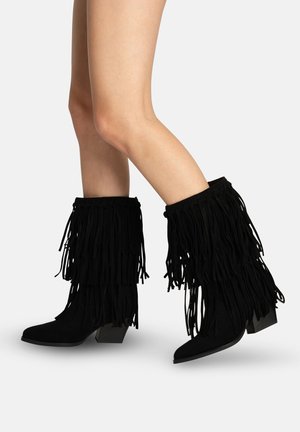 Piernas con botas negras de ante hasta la mitad de la pantorrilla, con flecos en capas y tacones cuadrados, sobre un fondo blanco.