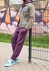 Džínová bunda světle béžové barvy, fialová mikina pod ní, fialové tepláky a modré tenisky Nike s zelenými a růžovými akcenty. Pozadí s graffiti.