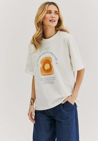 Bílá tričko s grafikou toastu se žlutým máslem. Text zní "Zlatá pauza v rušném světě" v modré barvě. Model má na sobě tmavé džíny.