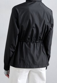 Svart lättviktsjacka med insamlad elastisk midja och muddar, bärs över ljusblå långärmad tröja och vita byxor, sedd bakifrån.