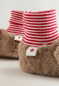 Pantoufles brunes en peluche avec un revers en tricot rayé rouge et blanc. Présente une petite étiquette blanche avec une étoile rouge sur le côté. Matériau doux et texturé.