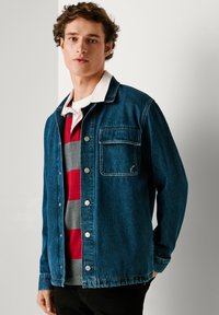 Giacca di jeans blu scuro con chiusura a bottoni, tasca sul petto singola e vestibilità comoda, indossata sopra un maglione a righe rosse e grigie.