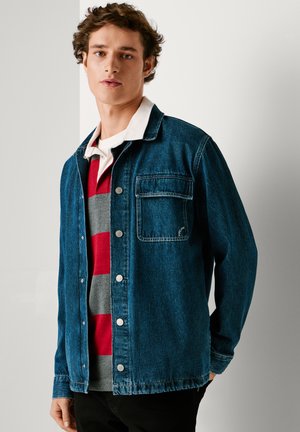 Tumed sineljakas nupuga esiosas, üheaastane rinna-tasku ja vabalt istuv lõige, kantud üle punase ja halli triibulise sviidri.