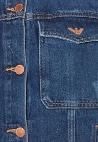 Džínová bunda ve tmavě modré barvě s měděnými knoflíky. Obsahuje náprsní kapsu a podpisové logo. Prošívané detaily přidávají texturu a zajímavost.