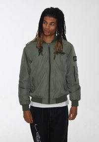 Giacca bomber verde oliva con polsini e fondo a coste, dotata di zip frontale, tasche laterali e un logo ricamato sul petto. Pantaloni neri.
