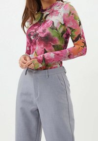 Halenka s dlouhým rukávem z průsvitného materiálu s barevným květinovým vzorem na bílém pozadí, v kombinaci se světle šedými kalhotami s předními kapsami.